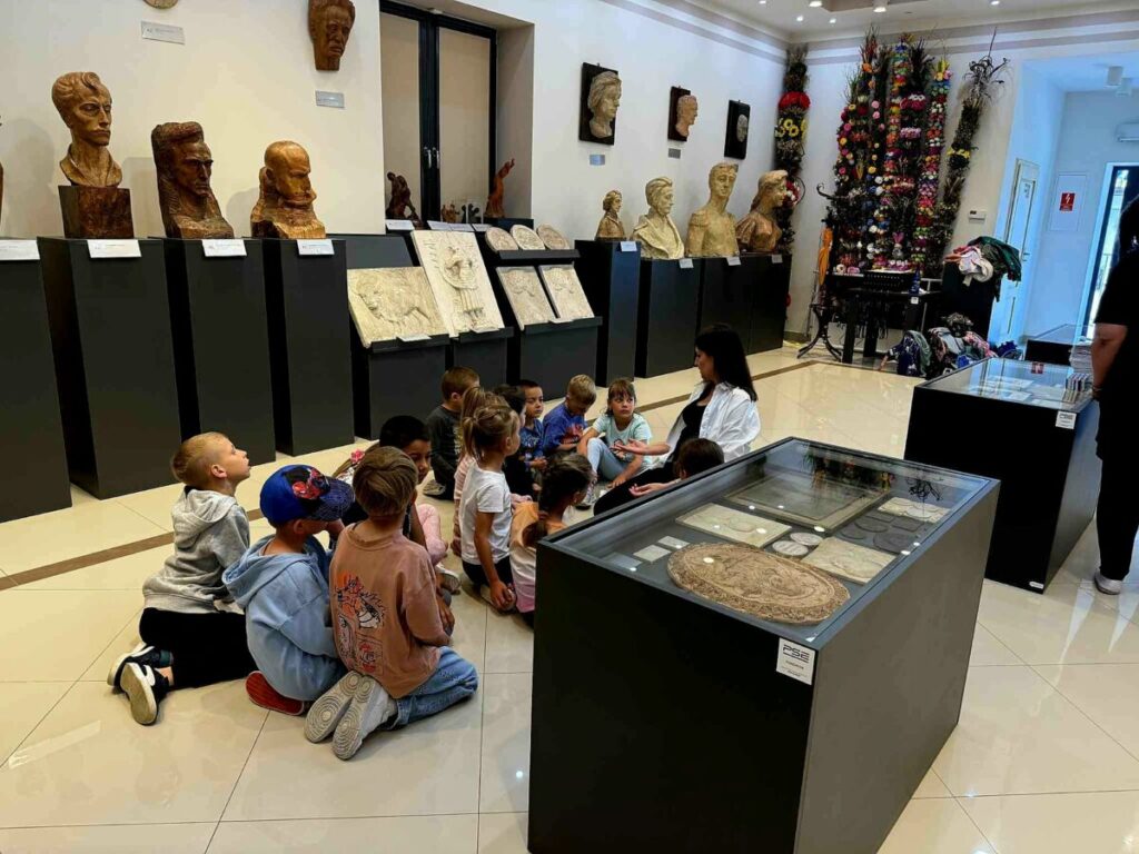 Grupa dzieci siedzi na podłodze w muzeum, oglądając wystawę rzeźb i płaskorzeźb. Ściany są jasne, a podłoga błyszcząca. Obok znajdują się szklane gabloty z eksponatami.