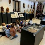 Grupa dzieci siedzi na podłodze w muzeum, oglądając wystawę rzeźb i płaskorzeźb. Ściany są jasne, a podłoga błyszcząca. Obok znajdują się szklane gabloty z eksponatami.