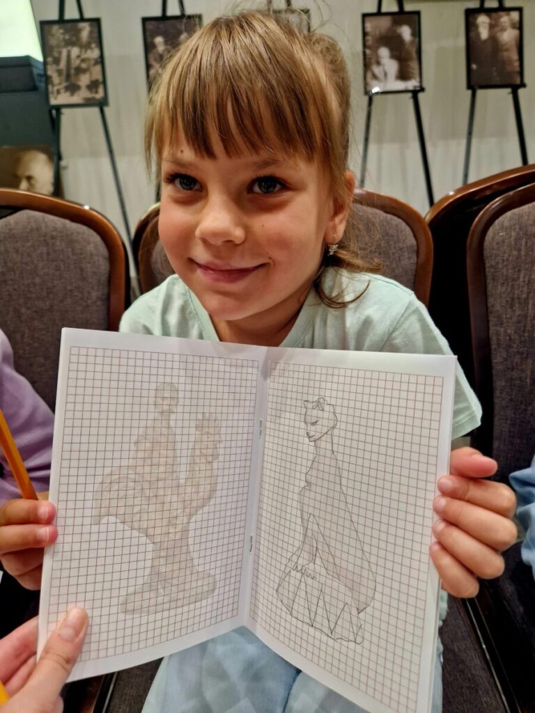 Dziewczynka trzyma szkicownik z rysunkami na kratkowanym papierze. Tło zdobią czarno-białe fotografie na stojakach. Ubrana jest w jasną bluzkę, a jej włosy są jasnobrązowe.