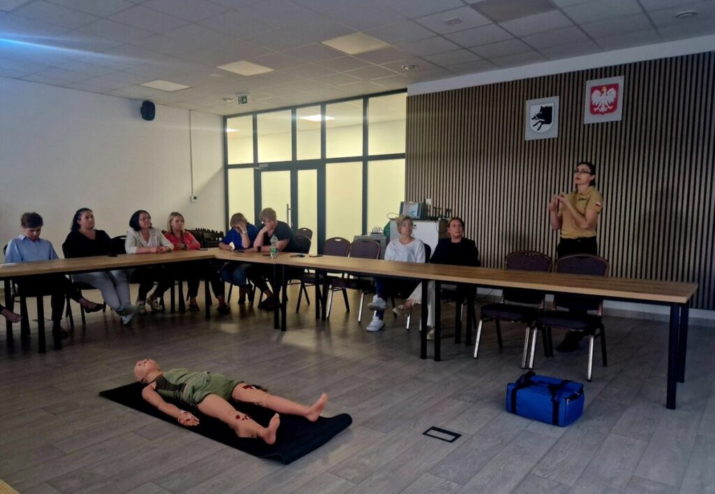 Grupa siedzi wokół stołu w sali konferencyjnej. Na środku manekin na czarnej macie. Kobieta prowadzi prezentację przy tablicy.