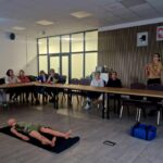 Grupa siedzi wokół stołu w sali konferencyjnej. Na środku manekin na czarnej macie. Kobieta prowadzi prezentację przy tablicy.