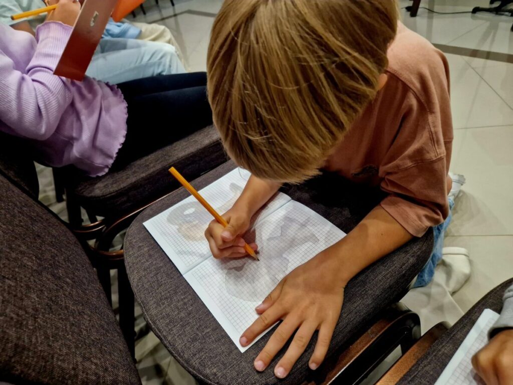 Dziecko rysuje ołówkiem na kartce w linie, siedząc na krześle. Bluzka różowa, tło jasne z ciemnymi krzesłami. Obok inne dziecko pisze w zeszycie.
