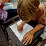 Dziecko rysuje ołówkiem na kartce w linie, siedząc na krześle. Bluzka różowa, tło jasne z ciemnymi krzesłami. Obok inne dziecko pisze w zeszycie.