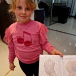 Dziewczynka w różowej bluzce trzyma kartkę z siatką i rysunkiem. W tle jasne wnętrze z wystawą. Podłoga błyszcząca, jasna.