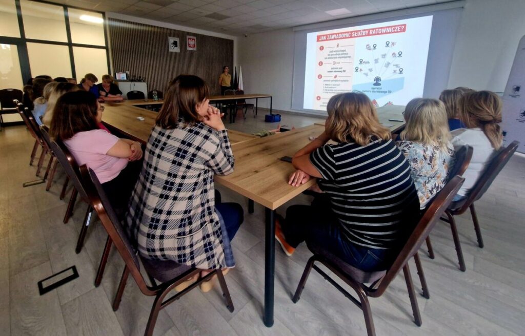 Grupa ludzi siedzi przy dużym stole i patrzy na ekran z prezentacją w jasnej sali. Na ekranie widoczny jest tekst o powiadamianiu służb ratunkowych. Kolory to głównie biel, brąz i szarość.