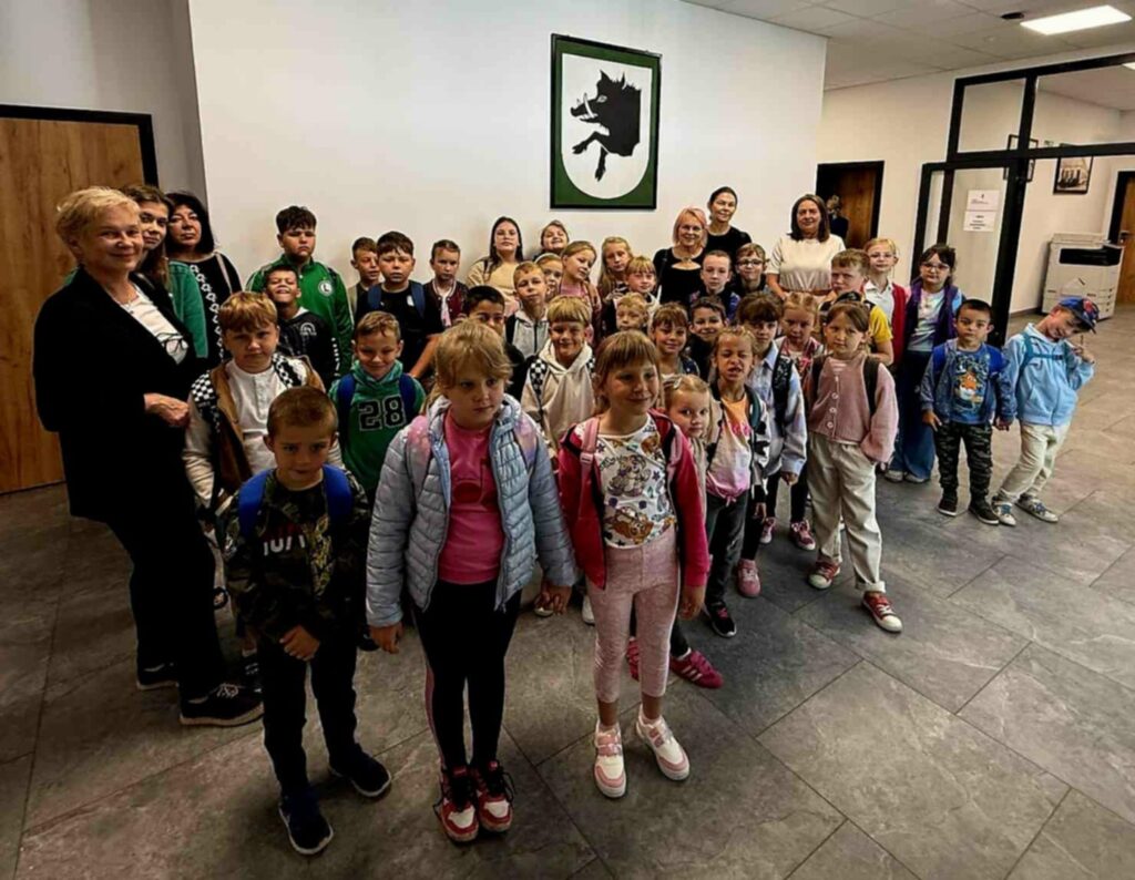 Grupa dzieci i dorosłych stoi w holu na tle białej ściany z zielonym obrazem. Dzieci mają kolorowe ubrania i plecaki. Podłoga jest ciemnoszara.