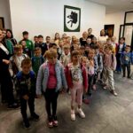 Grupa dzieci i dorosłych stoi w holu na tle białej ściany z zielonym obrazem. Dzieci mają kolorowe ubrania i plecaki. Podłoga jest ciemnoszara.