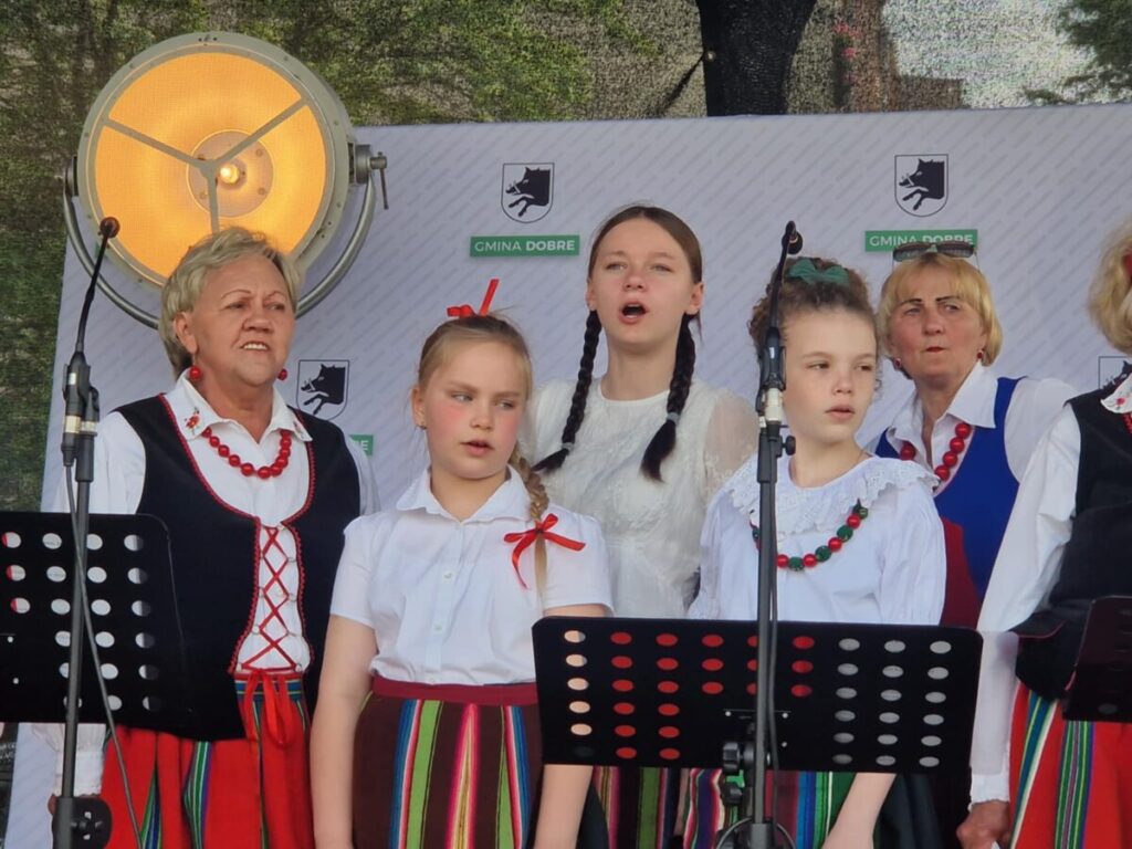 Grupa osób w strojach ludowych śpiewa na scenie. Tło z logo gminy i dużym reflektorem. Przeważają kolory czerwony, biały i zielony.