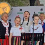 Grupa osób w strojach ludowych śpiewa na scenie. Tło z logo gminy i dużym reflektorem. Przeważają kolory czerwony, biały i zielony.