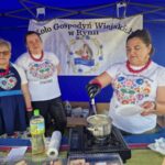 Trzy kobiety w ludowych strojach stoją przy stoisku z niebieskim tłem. Jedna z nich gotuje w garnku. Widoczny jest baner Koła Gospodyń Wiejskich.