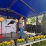 Osoba przemawia na scenie z niebieskim zadaszeniem. Tło z napisem Gmina Dobre. Przód sceny ozdobiony żółtymi kwiatami.