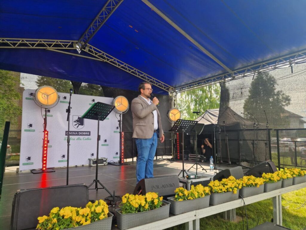 Na scenie pod niebieskim namiotem stoi osoba w jasnej marynarce i dżinsach, przemawiając do mikrofonu. W tle widoczne są reflektory i baner z napisem GMINA DOBRE. Przed sceną znajdują się żółte kwiaty.
