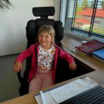 Dziewczynka w różowej bluzie siedzi na czarnym fotelu przy biurku. W tle są biurowe okno i roślina. Na biurku leży stos dokumentów.