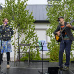 Na scenie stoją kobieta z mikrofonem i mężczyzna grający na gitarze. W tle są białe budynki i zielone drzewa. Skromna aranżacja sceny z krzesłem i pulpitem na nuty.