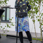 Kobieta z mikrofonem w kolorowym ubraniu stoi na scenie. Tło to białe ściany i zielone drzewa. Szenę uzupełniają ciemne płyty podłogowe.