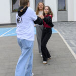 Trzy osoby bawią się na betonowym chodniku przed białym budynkiem. Noszą kolorowe ubrania, w tle widać okna. Jedna osoba ma ciasteczko w ustach.