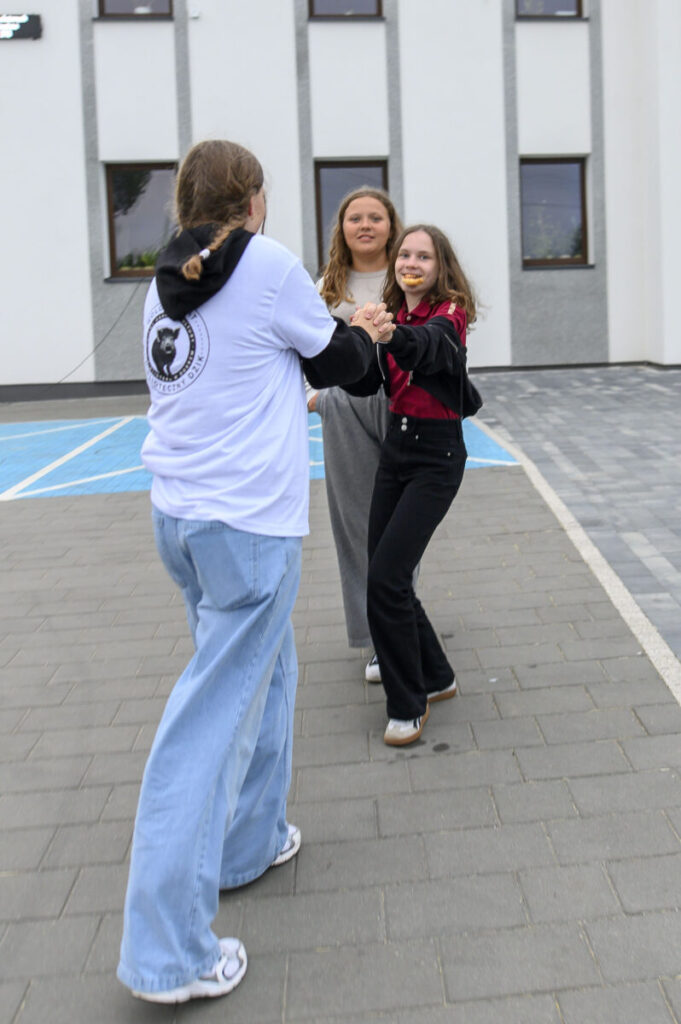 Trzy osoby bawią się na betonowym chodniku przed białym budynkiem. Noszą kolorowe ubrania, w tle widać okna. Jedna osoba ma ciasteczko w ustach.