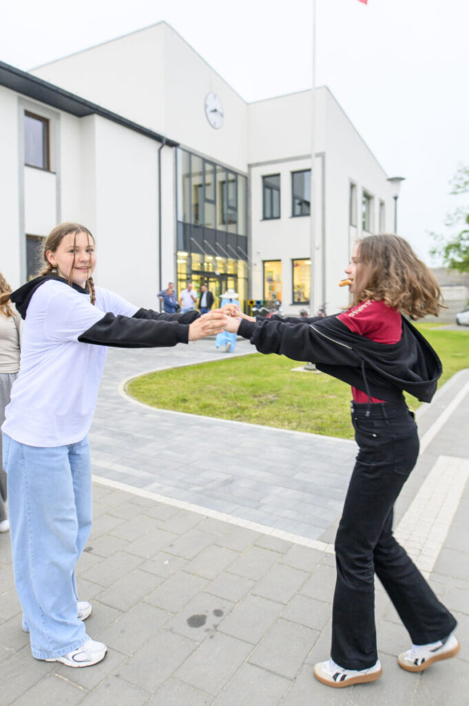 Dwie osoby tańczą przed nowoczesnym białym budynkiem. Dziewczyna w białej bluzie, druga w czerwonej koszulce. W tle zieleń i chodnik.