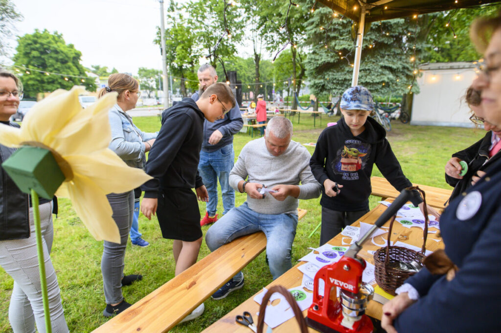 Grupa ludzi na pikniku tworzy przypinki na drewnianym stole. W tle zielone drzewa i światełka. W lewym rogu żółty, duży kwiat.