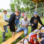 Grupa ludzi na pikniku tworzy przypinki na drewnianym stole. W tle zielone drzewa i światełka. W lewym rogu żółty, duży kwiat.