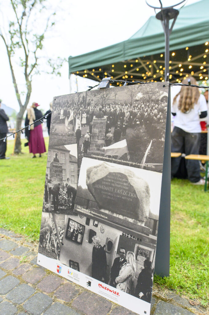 Plakat z czarno-białymi fotografiami historycznymi stoi na zielonej trawie. W tle namiot z lampkami i ludzie. Obok drzewo.