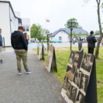 Osoby oglądają wystawę plenerową z czarno-białymi zdjęciami. Tło to budynki oraz flaga Polski. Chodnik w szarych tonacjach.