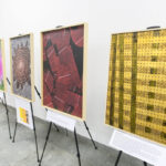 Wystawa z pięcioma obrazami na sztalugach w galerii. Obrazy mają jaskrawe kolory: różowy, czerwony, zielony i żółty. Tło białe, posadzka szara.