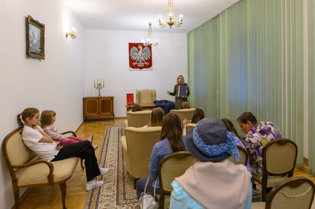Sala z grupą słuchaczy, kobieta prowadzi wykład. Jasne ściany, żyrandole, zielone zasłony. Obraz orła na ścianie.