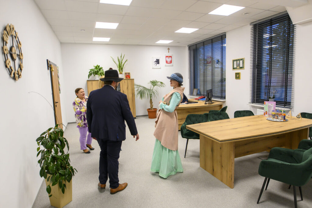 Pomieszczenie biurowe z drewnianymi stołami i zielonymi krzesłami. Trzy osoby rozmawiają, ubrania w kolorach fioletowych i miętowych. Na ścianie zegar, rośliny i dekoracje.