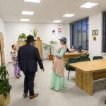 Pomieszczenie biurowe z drewnianymi stołami i zielonymi krzesłami. Trzy osoby rozmawiają, ubrania w kolorach fioletowych i miętowych. Na ścianie zegar, rośliny i dekoracje.