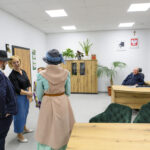 Cztery osoby w biurze, dwie w eleganckich kapeluszach. Tło w jasnych barwach, z roślinami i drewnianymi meblami. Na ścianie widać godło i plakat.