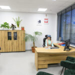 Biuro z drewnianym biurkiem i szafką, zielone rośliny, kobieta w kapeluszu przy komputerze, druga kobieta w kolorowym stroju.