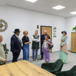 Grupa ludzi stoi w jasnym pomieszczeniu z drewnianymi meblami i roślinami. Na ścianie wisi duży zegar. Osoby są różnorodnie ubrane.