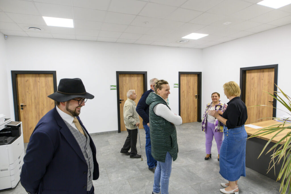 Grupa ludzi rozmawia w jasnym pomieszczeniu z szarymi kafelkami. Drzwi mają drewniane wykończenie. Z lewej strony stoi mężczyzna w kapeluszu i garniturze.