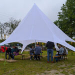 Pod białym namiotem na trawie siedzą ludzie, odpoczywając na drewnianych ławkach. Otacza ich zielone, drzewiaste tło. Na pierwszym planie widoczne są kolorowe poduszki.