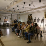 Grupa osób siedzi na krzesłach w jasnym pomieszczeniu z wystawą zdjęć na sztalugach. Tło jest białe z obrazami i delikatnym oświetleniem. Atmosfera przypomina wykład lub prezentację.