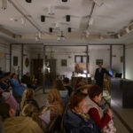 Grupa ludzi siedzi w oświetlonym pomieszczeniu. W tle mężczyzna w garniturze prowadzi prezentację. Ściany są białe z dekoracjami.