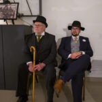 Dwóch mężczyzn w eleganckich garniturach i kapeluszach siedzi na krzesłach; jeden trzyma żółtą laskę. Tło białe, obok fotografia i laska w gablocie. Mają skupiony wyraz twarzy.