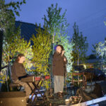 Muzycy występują na scenie na zewnątrz, w otoczeniu drzew i świateł. Niebieskie niebo zmierzchu stanowi tło. Wokalistka i klawiszowiec w ciemnych ubraniach.