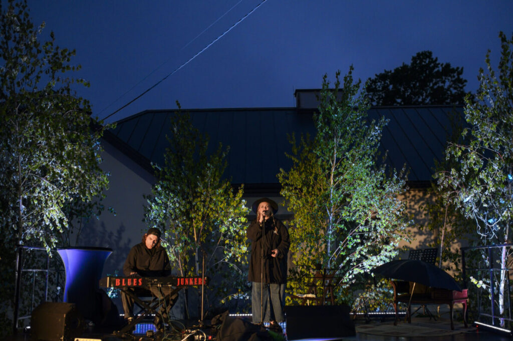 Koncert na świeżym powietrzu, wieczorem. Dwoje muzyków na scenie, wokół zielone drzewa. Niebieskie niebo w tle.