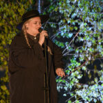 Osoba w czarnym stroju i kapeluszu śpiewa do mikrofonu. W tle zielone liście na drzewie. Scena oświetlona miękkim światłem.