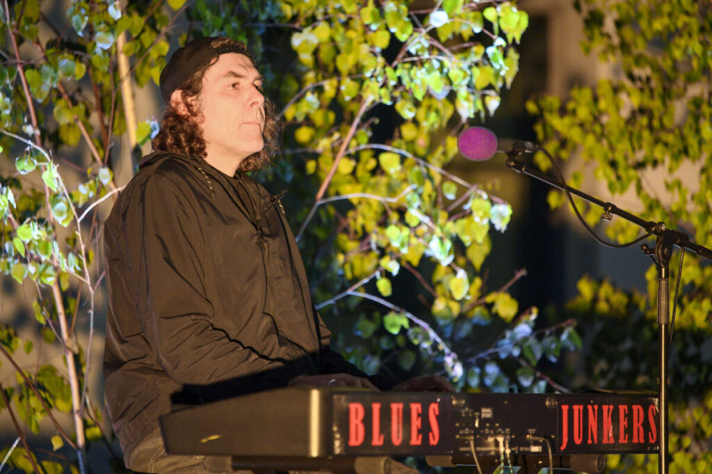 Osoba siedzi przy keyboardzie z napisem Blues Junkers. Ma na sobie ciemne ubranie i czapkę. W tle oświetlone drzewa.