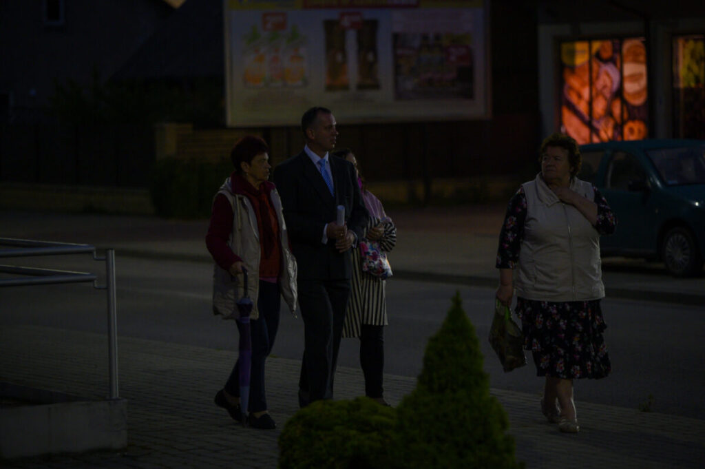 Grupa ludzi spaceruje po ciemnej ulicy, w tle billboard i zaparkowany samochód. Osoby są ciepło ubrane. Tło jest rozmyte.