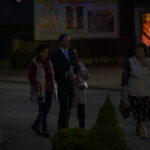 Grupa ludzi spaceruje po ciemnej ulicy, w tle billboard i zaparkowany samochód. Osoby są ciepło ubrane. Tło jest rozmyte.