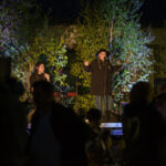 Koncert na zewnątrz z wokalistką w kapeluszu i muzykiem na klawiszach, tło z podświetlonymi, zielonymi drzewami.