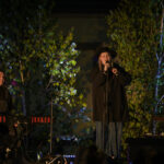 Koncert na świeżym powietrzu z dwoma muzykami na scenie. Na pierwszym planie klawisze i mikrofon, w tle zielone drzewa. Scena oświetlona na niebiesko.