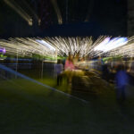 Rozmyte światła na ciemnym tle tworzą dynamiczny efekt linii. Przód ozdobiony świetlistymi paskami w ciepłych tonach. W oddali niewyraźne postacie.