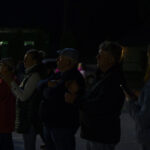 Grupa osób stoi na zewnątrz wieczorem. Mają założone ciepłe ubrania, w tym czapki i kurtki. Tło jest ciemne, subtelnie oświetlone.