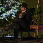 Mężczyzna w ciemnym garniturze i kapeluszu siedzi na scenie. Obok niego stolik z telefonem. W tle zielone liście na gałęziach.