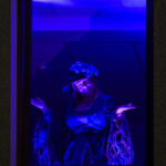 Portret osoby w kapeluszu, oświetlonej na niebiesko-fioletowo, z ciemnym tłem. Osoba ma rozłożone ręce. Zdjęcie w ramce.